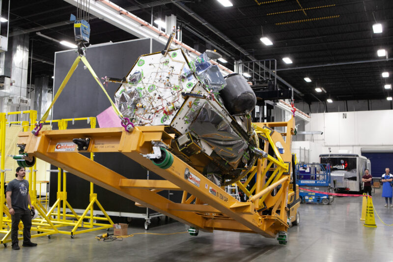 Silver foil-covered spacecraft lying on its side on a yellow lift jack while a worker, tiny by comparison, watches.
