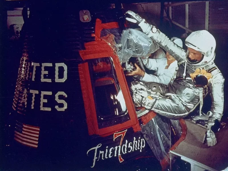 Man in silver suit writhing feet first into small space capsule with Friendship 7 written on the side.