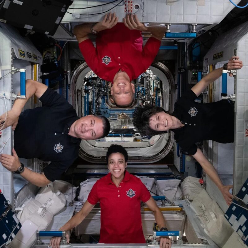 4 grinning people on 4 different sides like a compass inside the International Space Station.