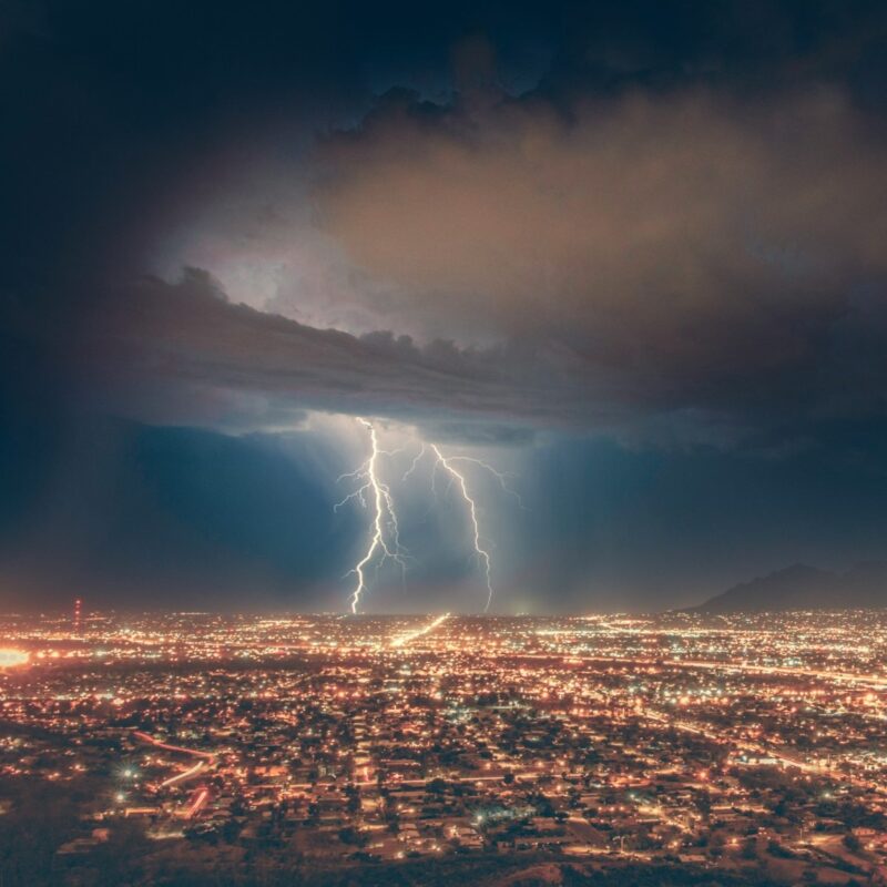 Lightning strikes from a dark, glowing cloud over the lights of a vast city.