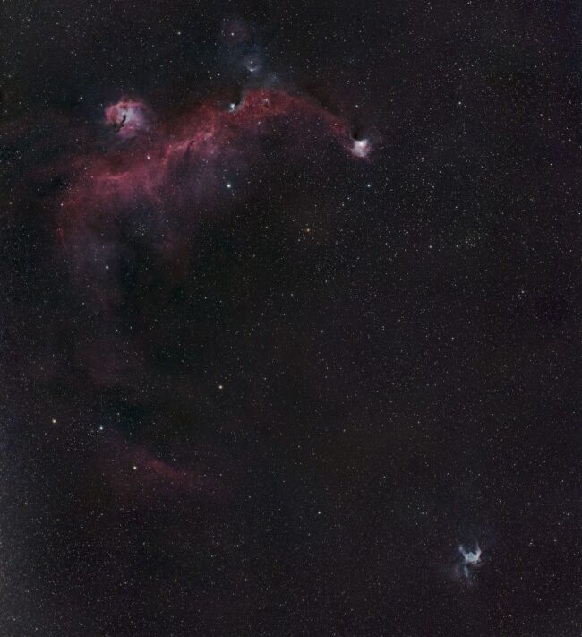 Wide, sweeping, fuzzy red nebula in a field of scattered stars, with another small blue one near it.
