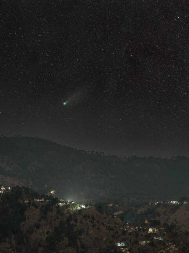 Mountain town at night with a comet that has a glowing head and long tail, in the sky above.