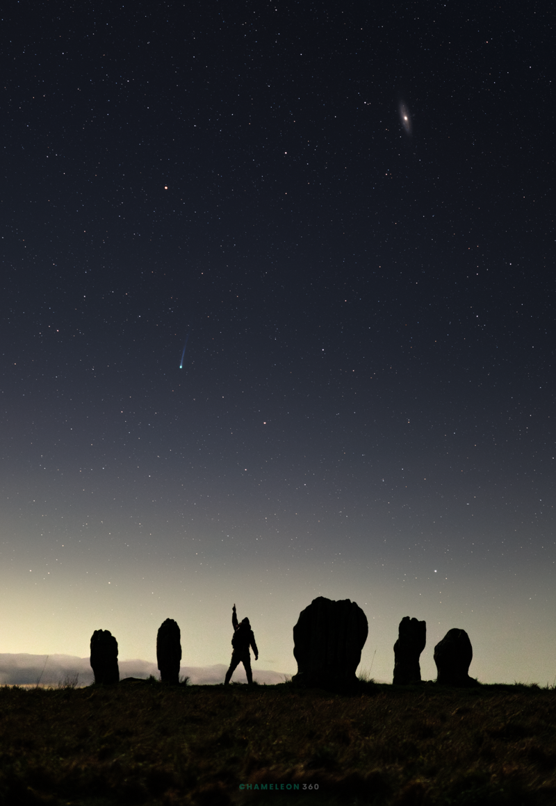 Five big rocks - the size of a human adult - in the foregrounds. A man stands among them and points to a starry sky.