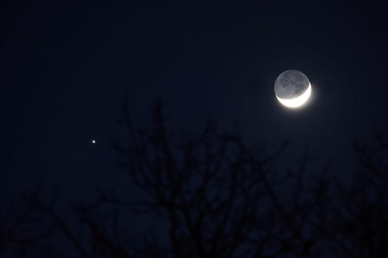 Bright crescent moon, with dark part glowing slightly. Dot with tiny dots for Jupiter and its moons.