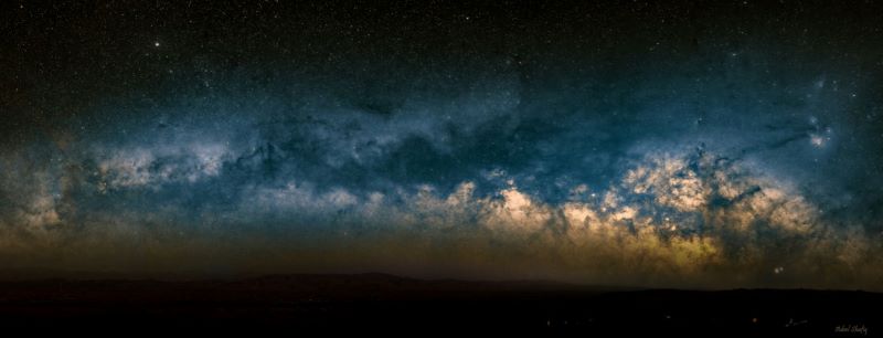 A dark cloud of dust with glowing stars behind stretching along the horizon.