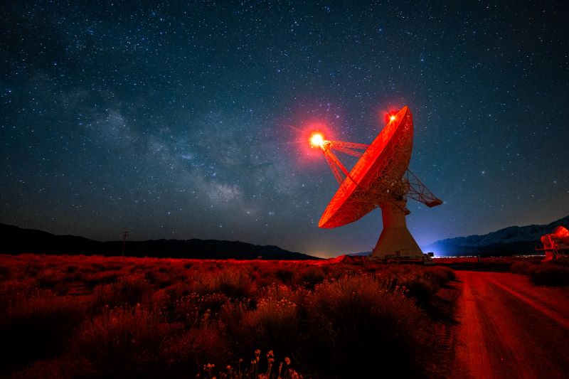 A radio dish glowing red with a band of stars behind it.