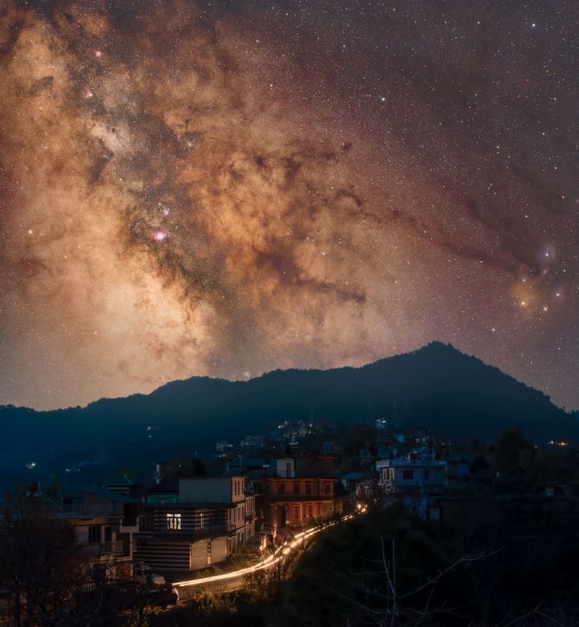 A bright glowing band of stars in the sky above a mountain and a town with some streaking car lights.