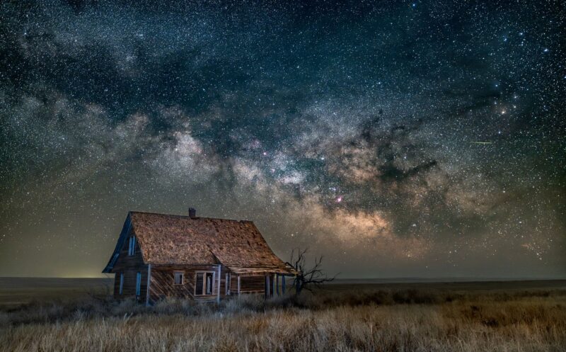 An old abandoned house on the prairie with a large, cloudy band of stars above.