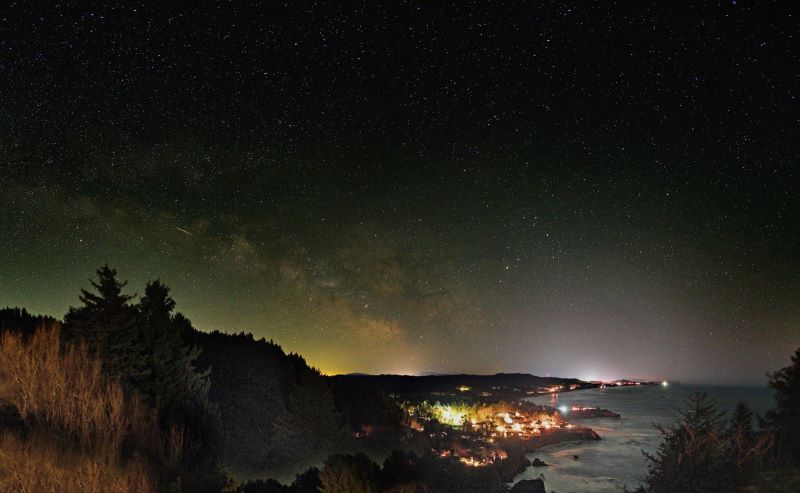 Una escena nocturna de una ciudad costera y una galaxia arqueada con un toque de luz en el lado izquierdo.