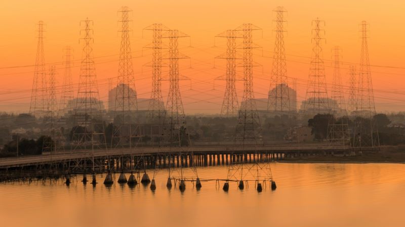 15 tall power towers with many lines between them against an orange sunset sky, water in the foreground.