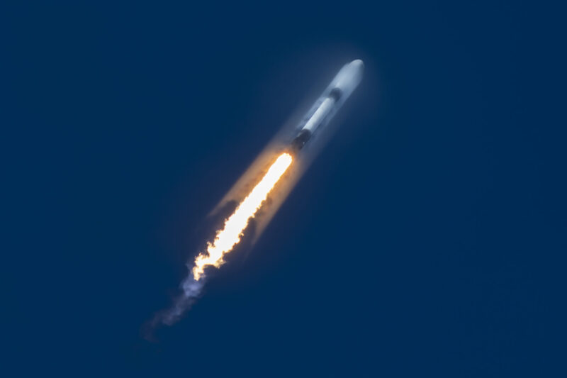 Image shows a rocket blasting through a deep blue sky with a jet of orange, yellow, and white propelling it upwards.