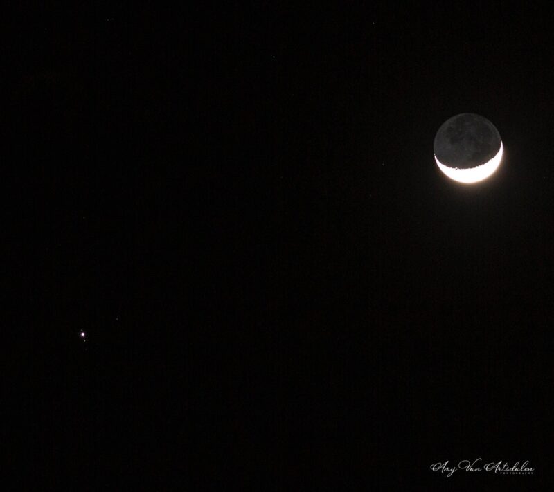 Bright crescent moon with dark part slightly glowing. Bright dot to its left with tiny dots in a line near it.