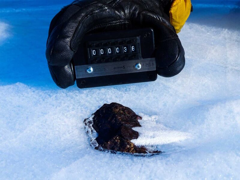 Antarctic meteorites: A partially submerged black rock in ice with a gloved hand holding a measurement tool.