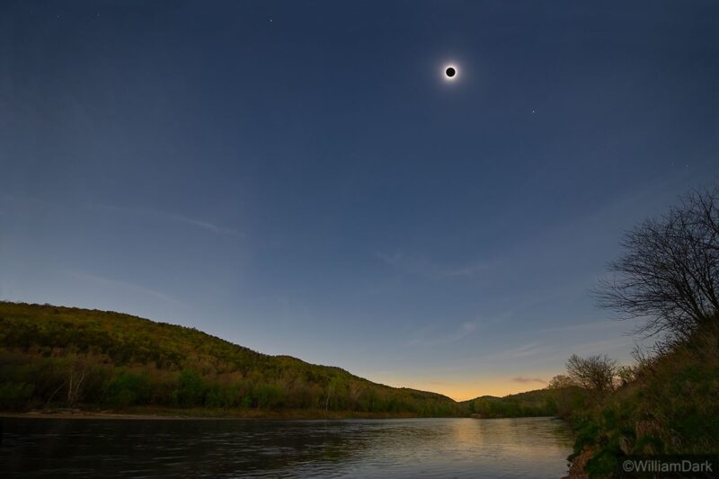 An eclipsed sun above a river with points of light to the upper left and lower right.