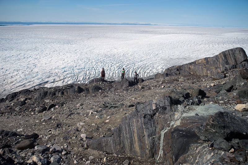 A brownish-gray rocky landscape with white ice fields in the distance. Several people are also seen in the distance.