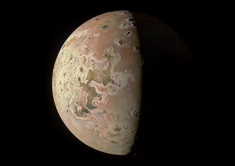 Io's volcanoes: Brownish, mottled and patchy planet-like body on black background.