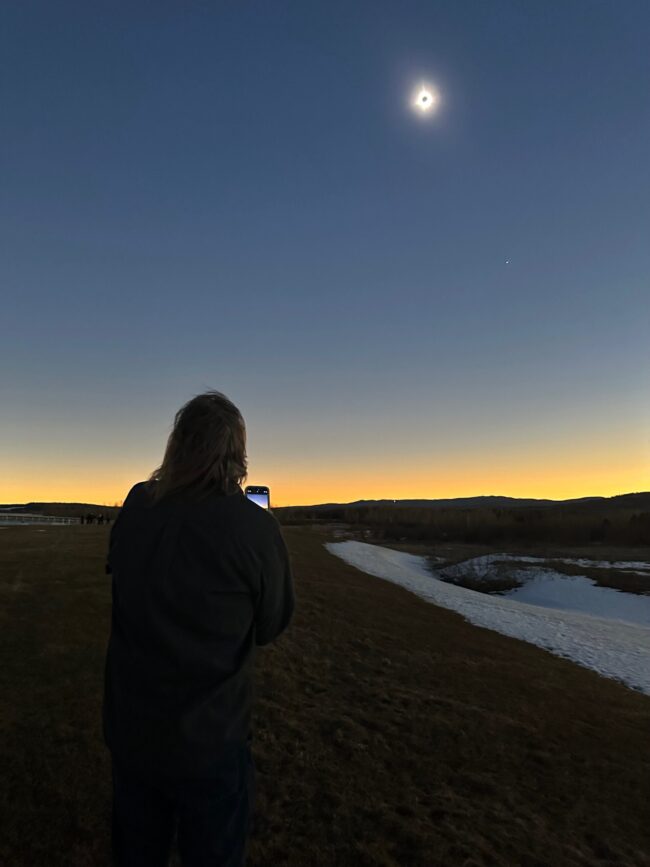 A woman stands in a sunset-like scene holding a smart phone and looking up at the eclipsed sun.