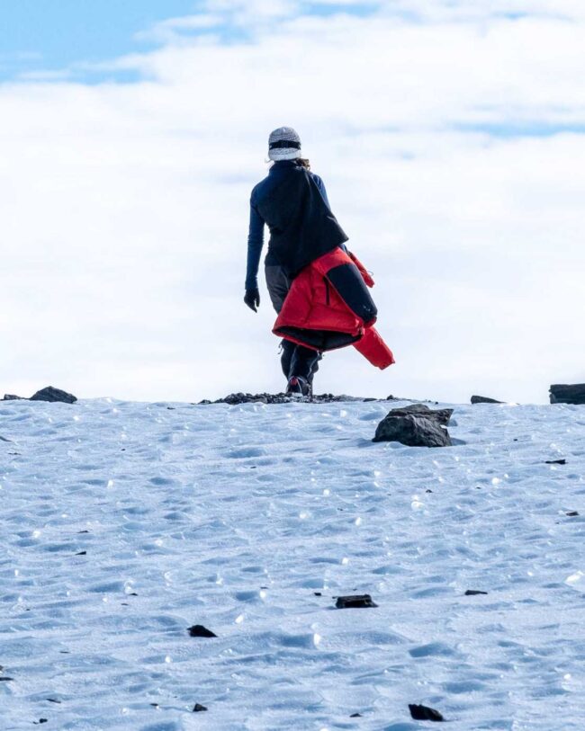 A woman carries a red jacket as she walks away from an ice field with black rocks strewn on top.