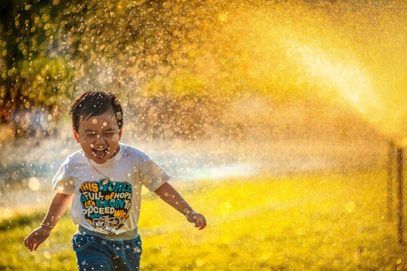 Un ragazzino corre attraverso gli irrigatori nel cortile e il sole splende attraverso l'acqua e sull'erba, rendendo dorato lo sfondo.