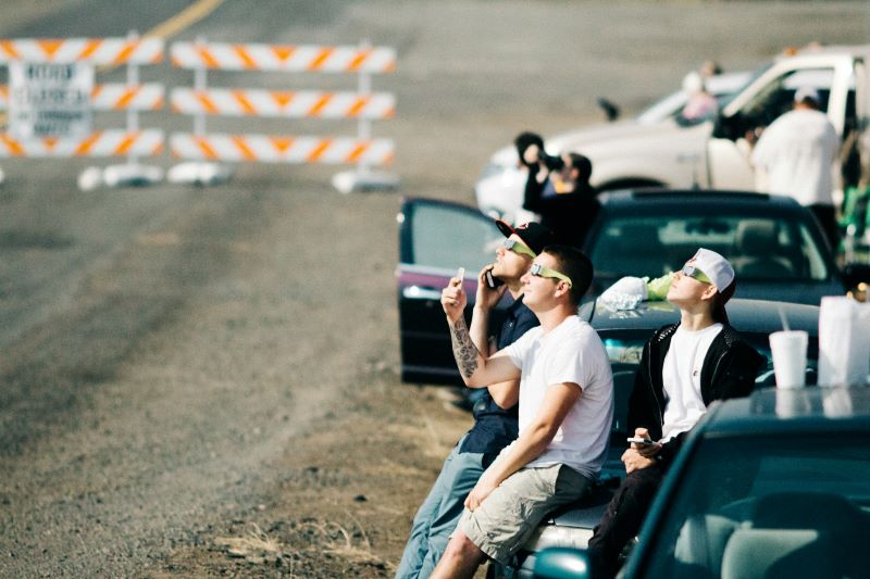 Some young men with eclipses glasses on leaning on their car and looking up at the sky.