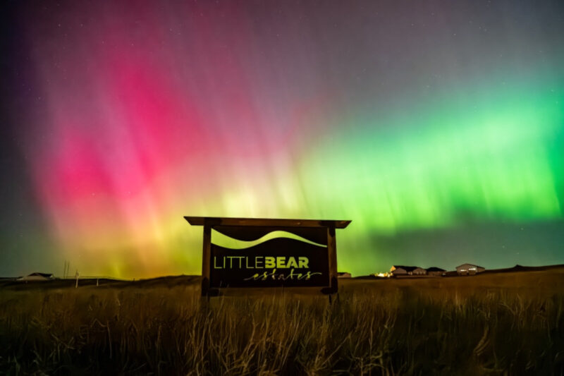 Brilliant red and green curtains of light high in the sky over a sign that says Little Bear Estates.