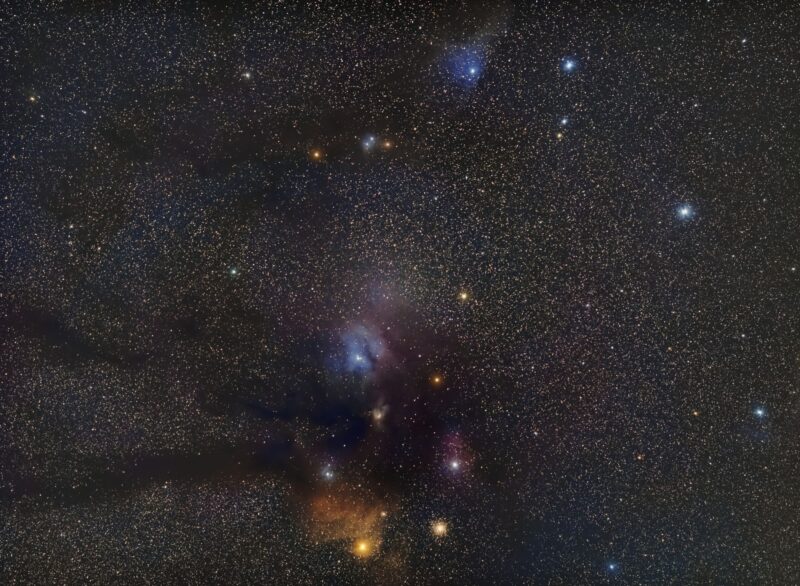 Small clouds of reddish, bluish and yellowish nebulosity over a multitude of background stars.