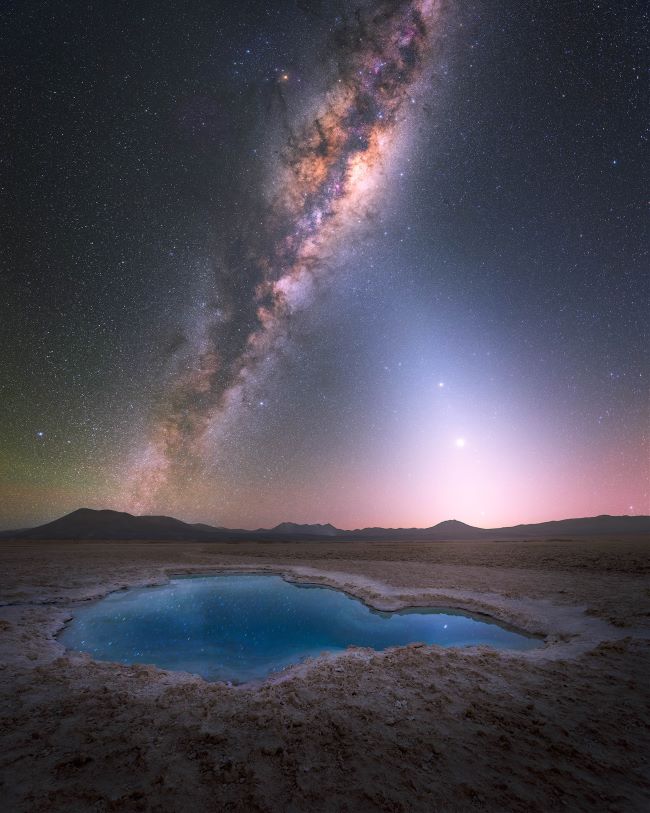 The Milky Way standing up in the sky with a white pyramidal shape to the right and a glowing blue pool in front.