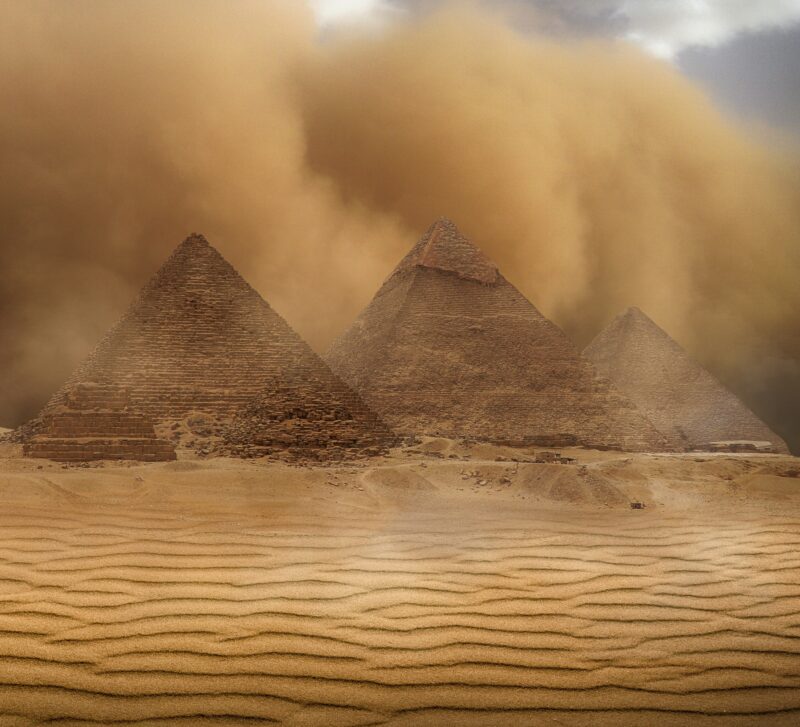 A desert with a sandstorm going on. There are 3 big pryamids in the center-right and 2 smaller ones on the left side.