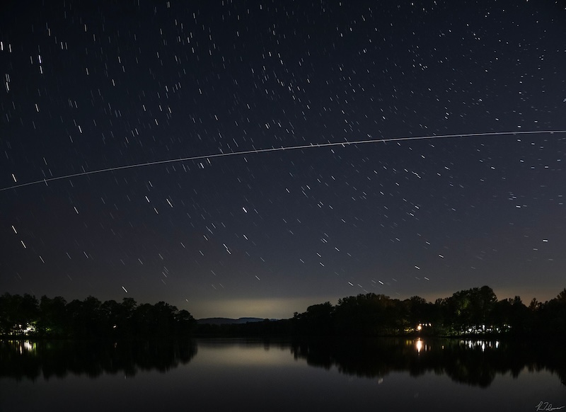 A white streak shoots across star trails in the sky. Over a body of water with trees and buildings in the distance.
