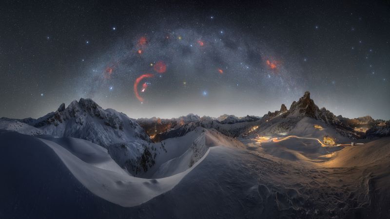 The Milky Way arching over a snowy, mountainous landscape with some roads and buildings in the mountains.
