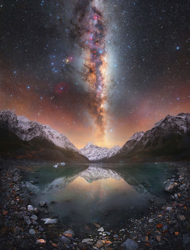 The Milky Way standing on end over a snow-capped mountain, with mountains on the edges and a lake in front.