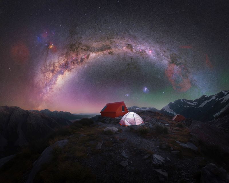 An arching Milky Way over a campsite in the mountains.