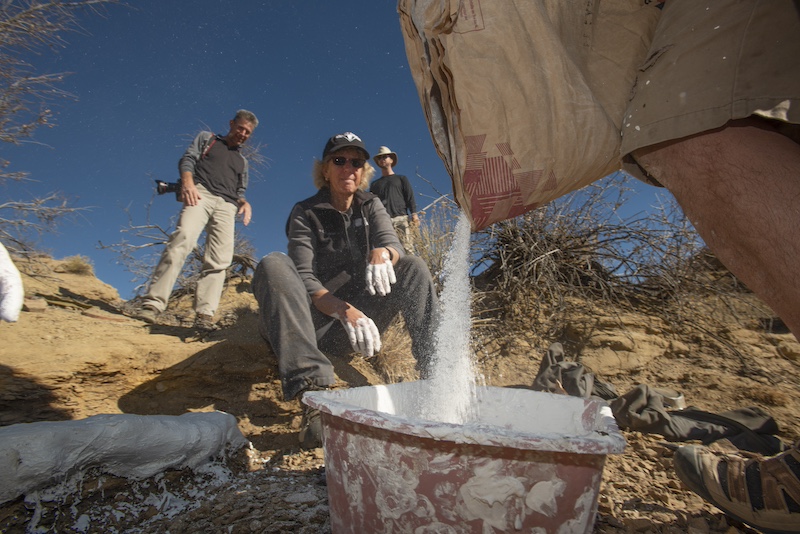 On rocky hillside, a closeup of white dust pouring from a bag into a tub, with scientists in the background under a blue sky.