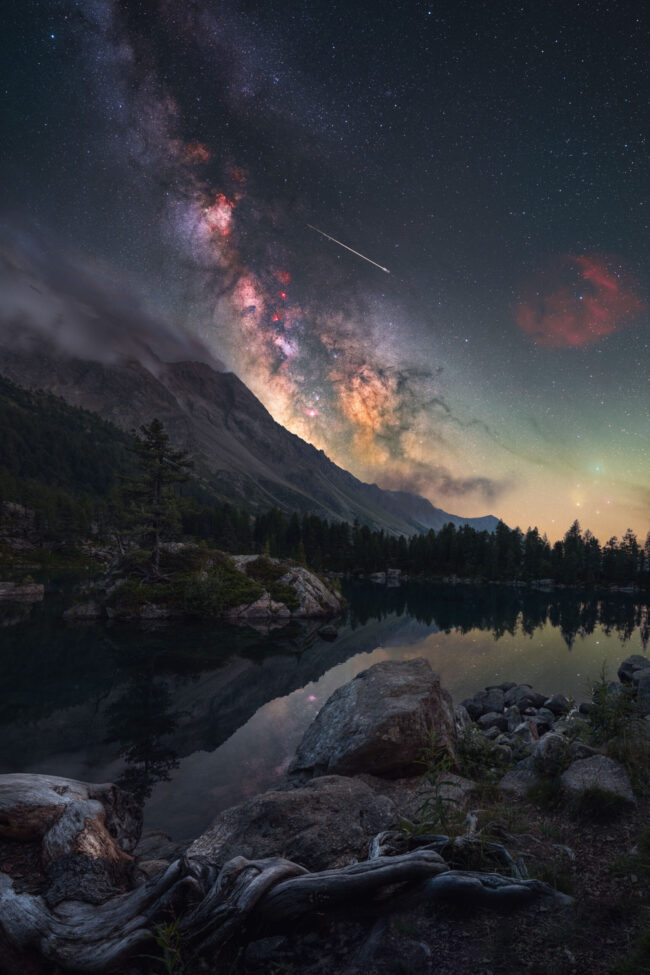 Mountainous terrain with brightly lit Milky Way behind and reflections in a pond.
