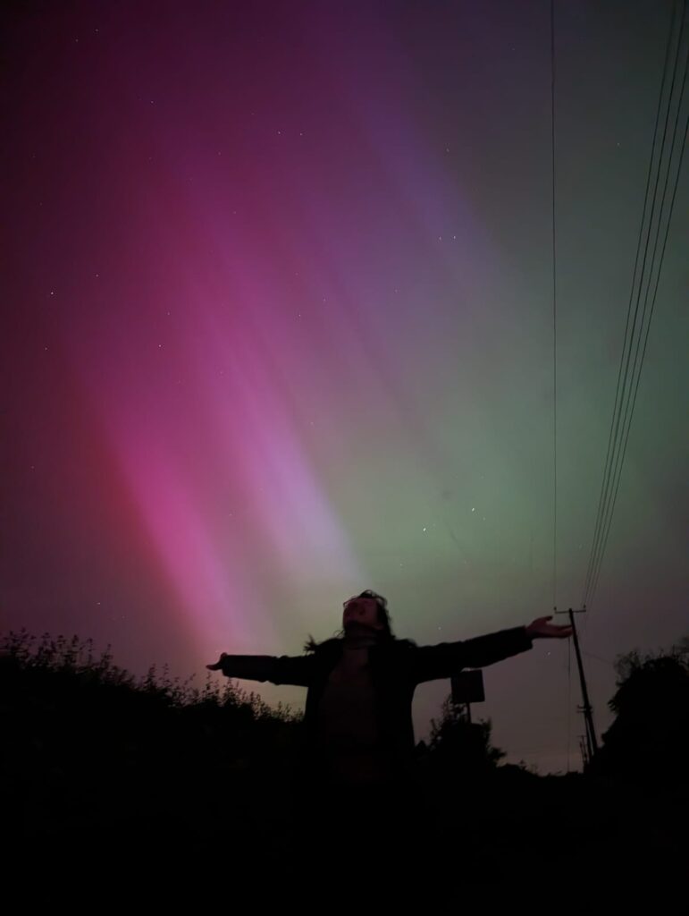 Pink, magenta, and green light stretches diagonally across the sky above the silhouette of somebody with their arms outstretched and their face lifted toward the sky.