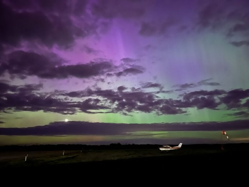 Streaks of purple and green light fill the sky behind scattered clouds above a small airplane on a dark field.