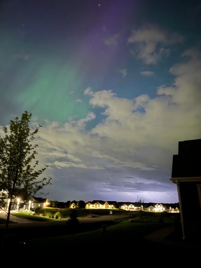 Clouds roll across a sky streaked with a green and purple sheen over a lit neighborhood street. In the distance, lightning flashes from the clouds.