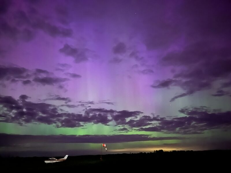Streaks of purple and green light fill the sky behind scattered clouds above a small airplane on a dark field.