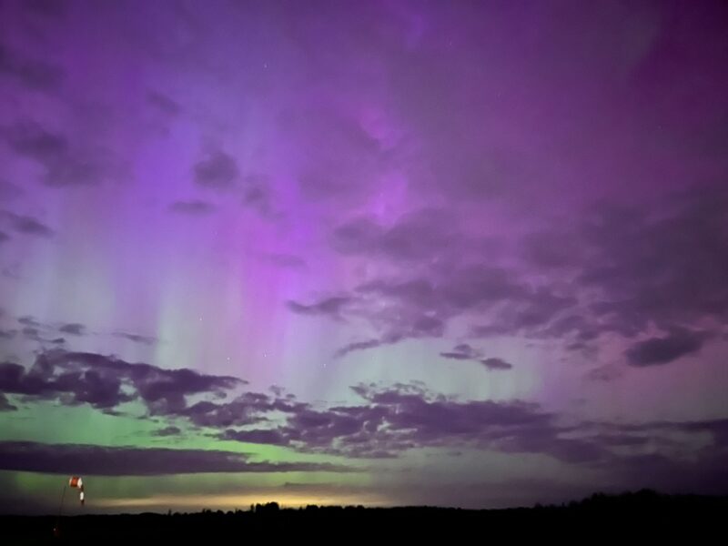 Streaks of purple and green light fill the sky behind scattered clouds above a dark field.