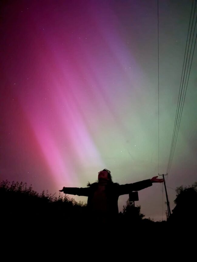 Pink, magenta, and green light stretches diagonally across the sky above the silhouette of somebody with their arms outstretched and their face lifted toward the sky.