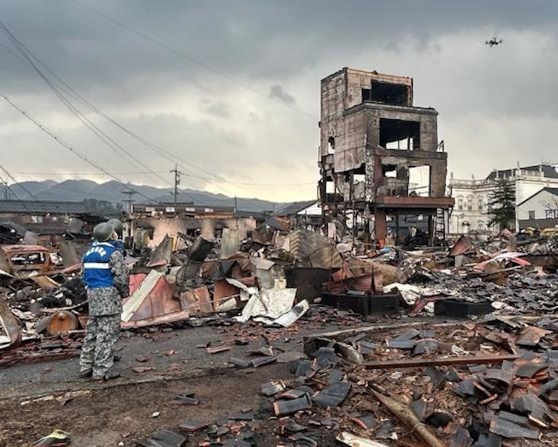 Earthquakes: Man standing looking at ruins of buildings, with drone flying overhead.