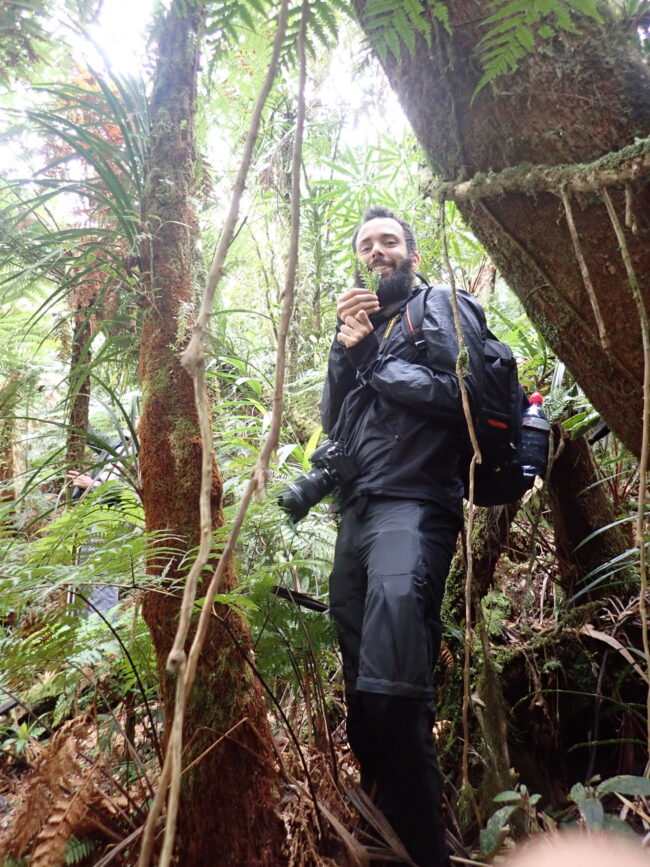 A man with dark facial hair and black clothes with backpack and camera holds up two small fronds.