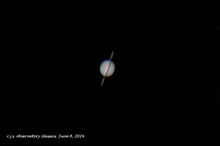 Saturn through a telescope shown as a grayish oval with the rings almost edge-on.