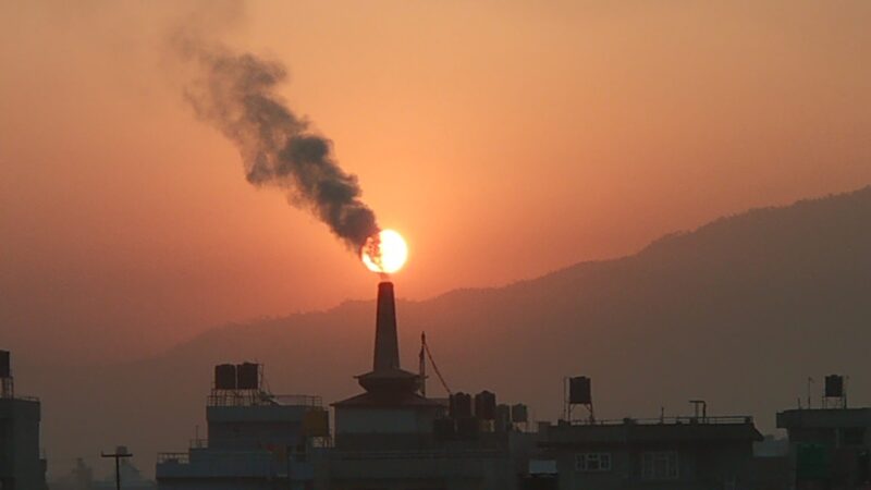 Smoke billowing from a tall factory smokestack. Background: Dim sun in smoggy orange sky.