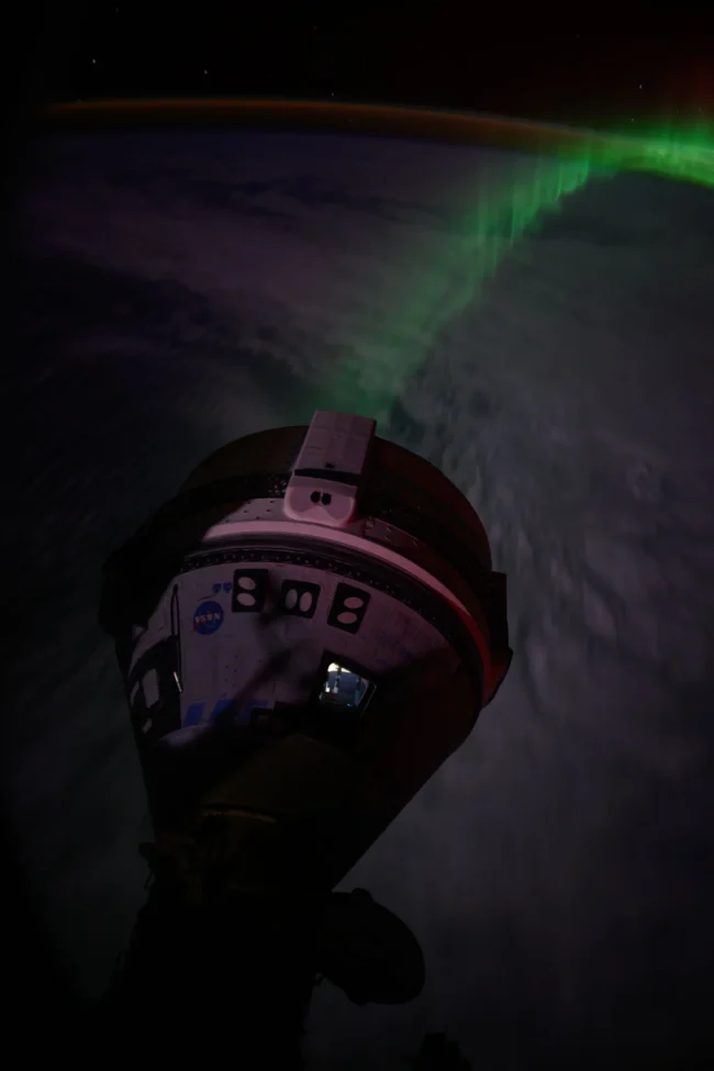Cloud-covered Earth at night from orbit. Conical Starliner crew capsule in partial shadow. Rainbow trail of auroras.