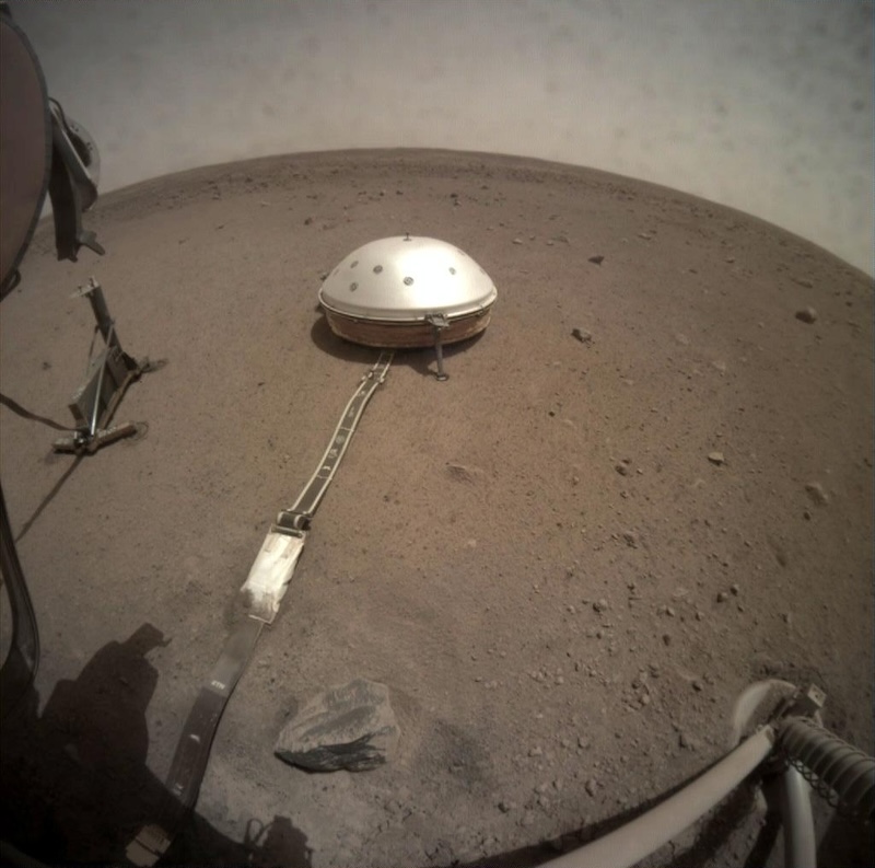 Small white metallic dome with long tether attached sitting on reddish dusty ground with small rocks and dusty sky.