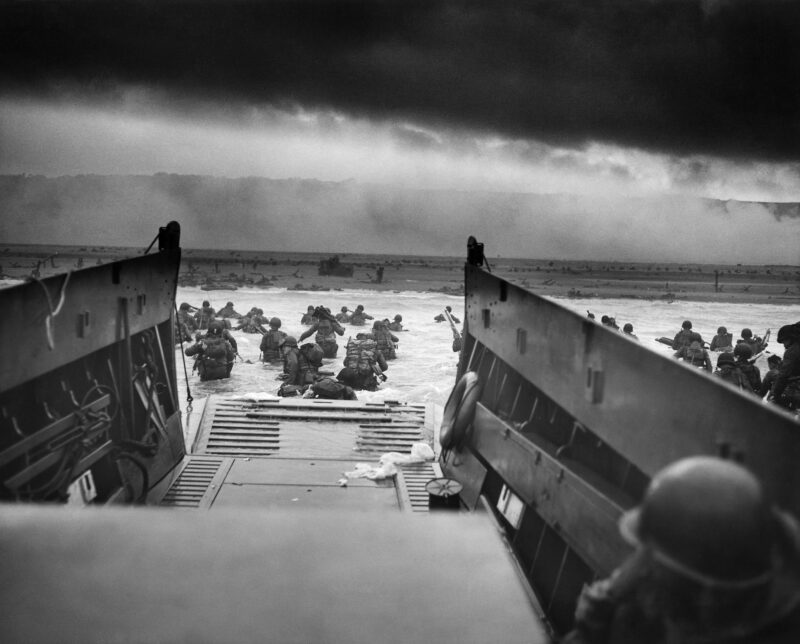 Black and white image. American troops in battle gear wade through the ocean from their landing craft. Dark band of clouds over fog. Landing craft in the foreground. D-Day