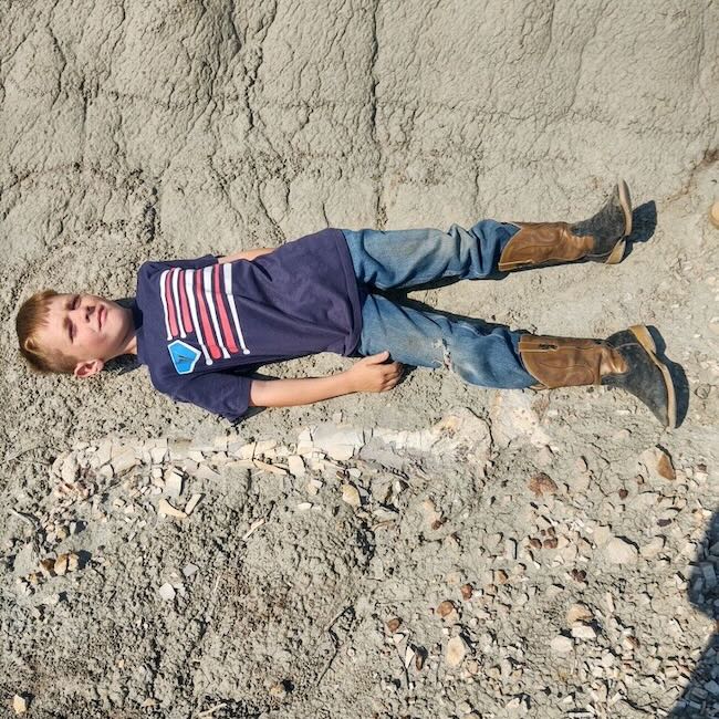A young boy lying next to a fossil legbone. The rock beneath him is grey. 
