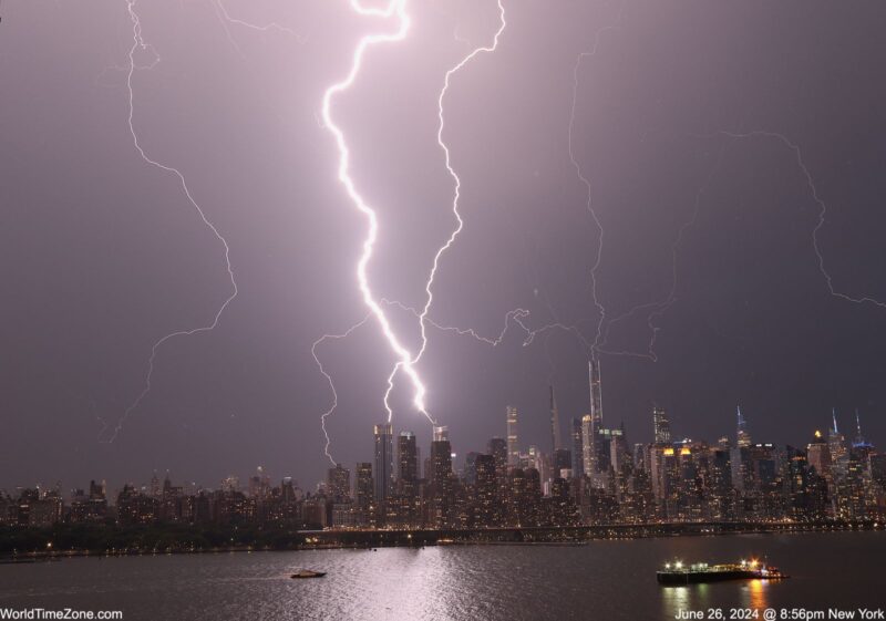 Lightning streaks from the sky to the ground, where there are many high buildings. The buildings are both illuminated by their yellow windows and the lighning strikes, also reflected in the water.