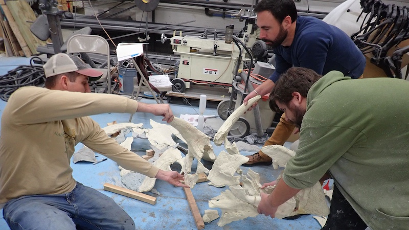 Three men in a lab bending over a table and working on the white casts of large bones.